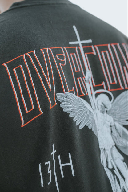 Close-up of the Overcome Premium Tee by 13th, a black shirt for young men. It features an angel graphic holding a spear and cross, with OVERCOME in red above and 13TH below, symbolizing strength through Gods word.