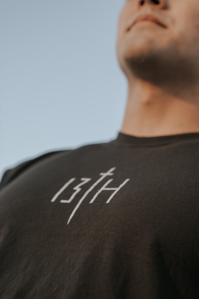 Close-up of a young man wearing the Overcome Premium Tee by 13th, a dark t-shirt featuring the brand name in white. His face is partially visible as he looks up at the clear blue sky, perhaps seeking inspiration to overcome challenges.