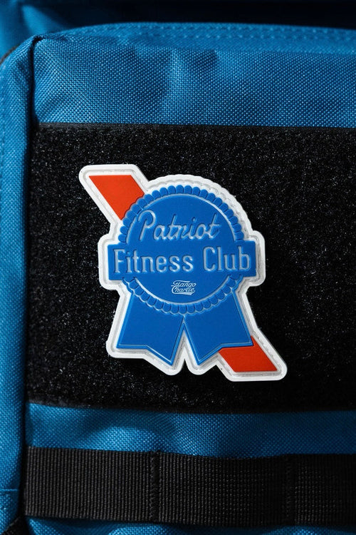 A blue backpack displays a 3-inch ribbon-shaped Patriots Fitness Club patch by Tango Charlie Apparel, in white and blue with a red stripe, attached to a black Velcro section.