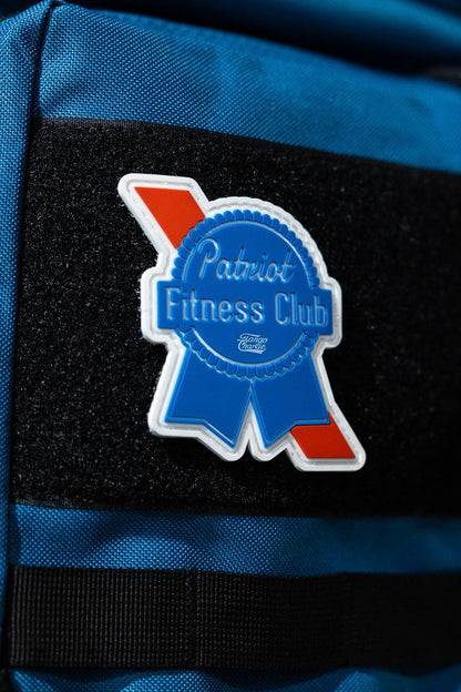 A Tango Charlie Apparel Patriots Fitness Club - Patch, shaped like a blue ribbon with red and white accents, is attached to the Velcro strip of a bright blue bag for a bold accessory.