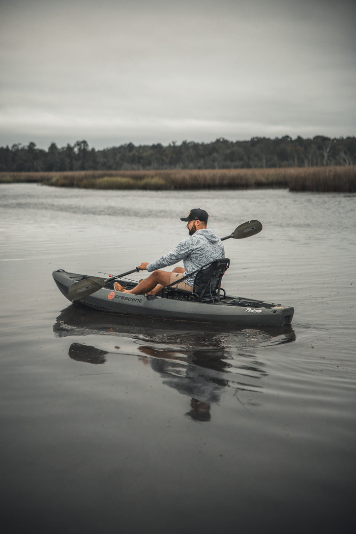 Wearing a Mucker Bucket Performance Hoodie - Ghost Camo and a cap, a person paddles a gray kayak on a calm river with grassy banks and trees under an overcast sky.