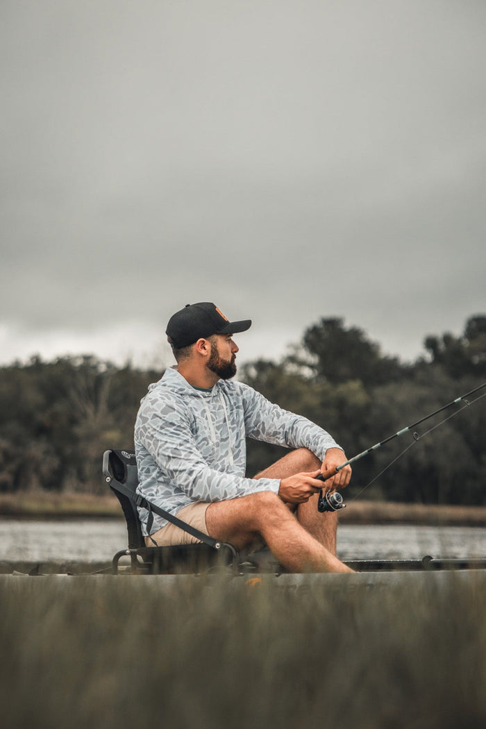 A man in the Mucker Bucket Performance Hoodie - Ghost Camo sits on a folding chair by the water, fishing rod in hand, with trees and a cloudy sky behind him.