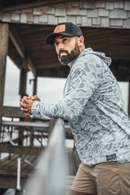 A bearded man in a Mucker Bucket Performance Hoodie - Ghost Camo and a black cap leans on a railing outdoors, gazing thoughtfully into the distance beneath a wooden shelter.