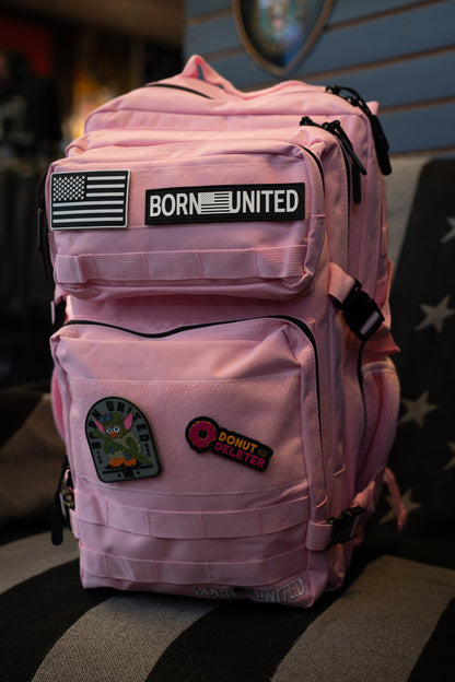 A pink tactical backpack with multiple zippered pockets, featuring a 'BORN UNITED' patch and other decorative patches, resting on a striped surface.