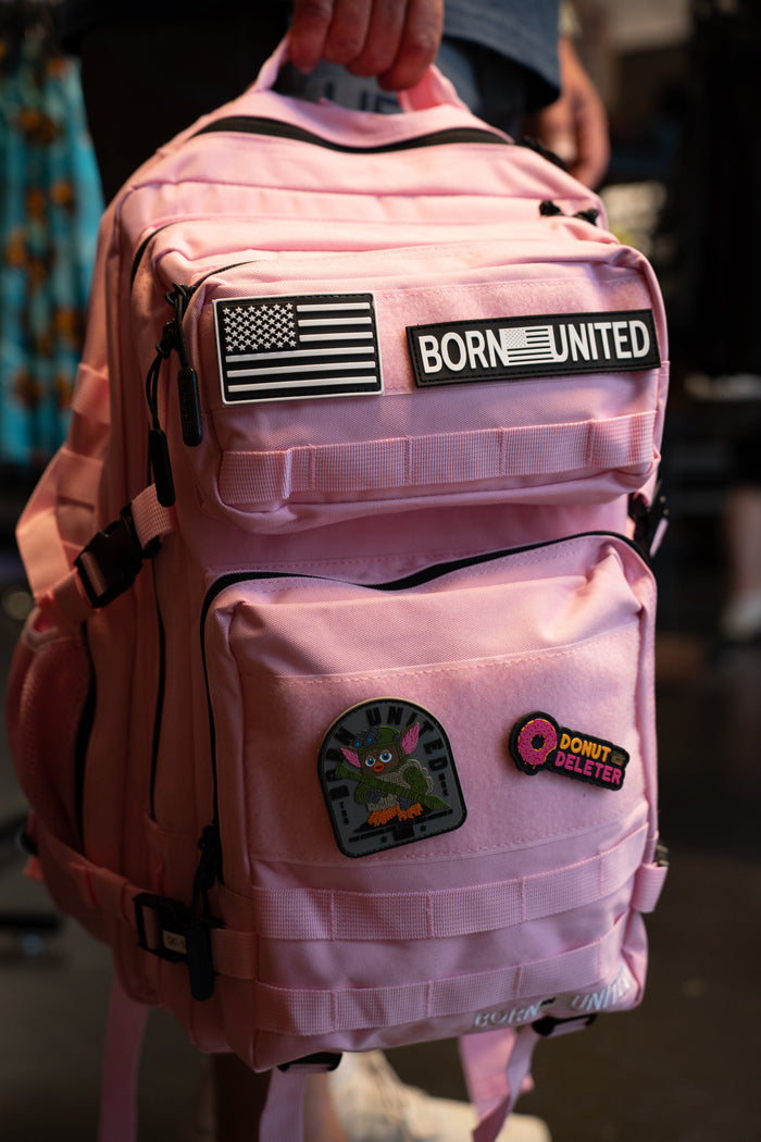 A pink backpack with black straps and multiple patches, including a 'BORN UNITED' patch and a bunny patch, being held by a person in a dark indoor setting.