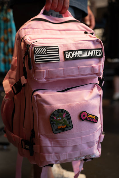 A pink backpack with black straps and multiple patches, including a 'BORN UNITED' patch and a bunny patch, being held by a person in a dark indoor setting.
