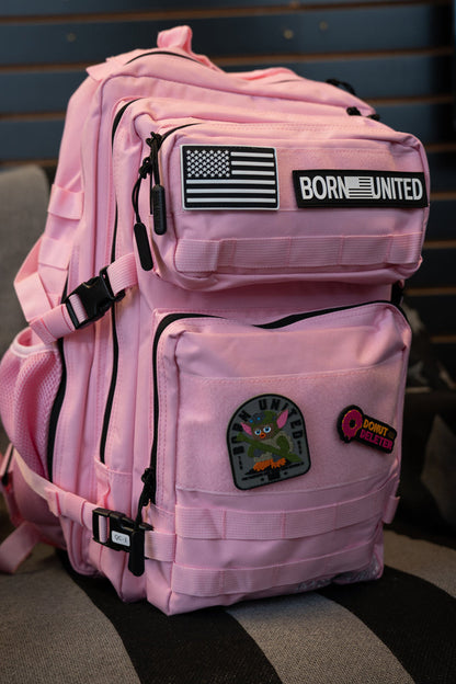 A pink tactical backpack with black zippers and multiple patches, including a 'BORN UNITED' patch, displayed on a striped gray and black seat.