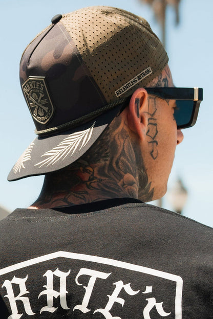 A person with neck tattoos wears a Pirates Life Camo Snapback by Relentless Betrayal, featuring a patch and RELENTLESS BETRAYAL on the strap. They have large sunglasses, a black shirt with bold white text, exuding adventure while keeping their face hidden.