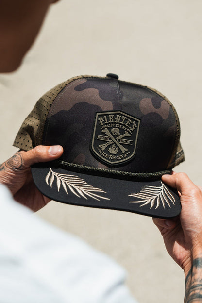 A person holds the Pirates Life Camo Snapback by Relentless Betrayal, a rugged camo baseball cap with crossed anchors and Pirate text patch, featuring perforated sides and embroidered palm leaves on the brim, capturing the adventurous spirit of a pirates life.