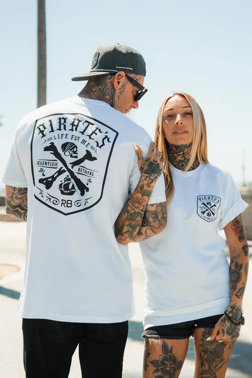 A tattooed man and woman wear matching Relentless Betrayal Pirate's Life White T-Shirts outdoors; he displays the back design, she faces forward—both radiate a confident, relaxed adventure vibe.