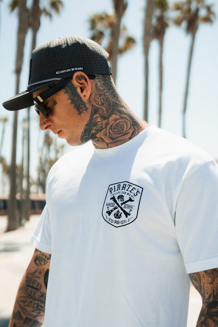 A tattooed man in sunglasses, a black cap, and the Relentless Betrayal Pirate's Life White T-Shirt stands among palm trees outdoors, embodying an adventurous lifestyle.