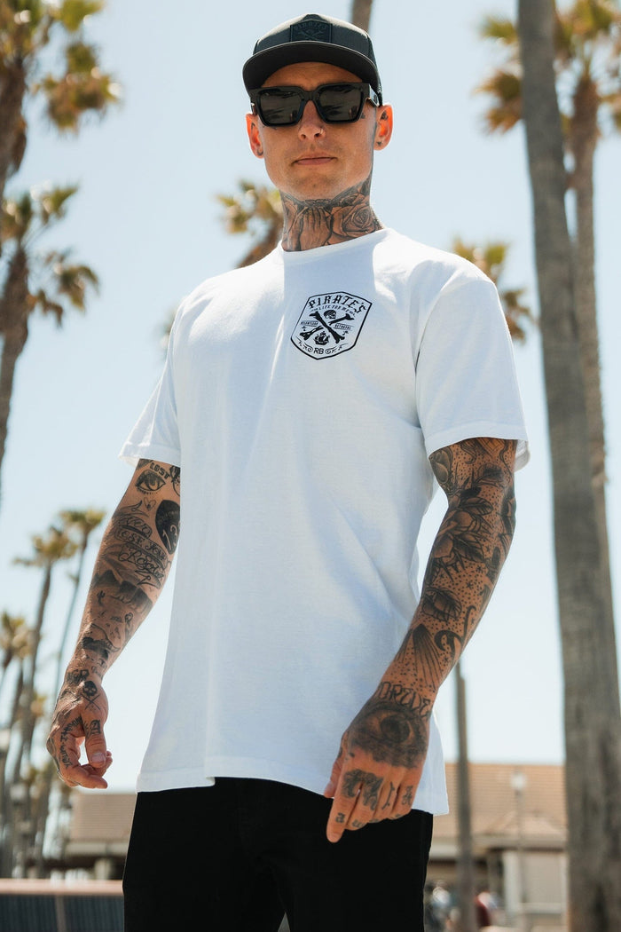 A tattooed man in a black cap and sunglasses wears the Relentless Betrayal Pirate's Life White T-Shirt outdoors, embracing adventure among palm trees and a clear sky.