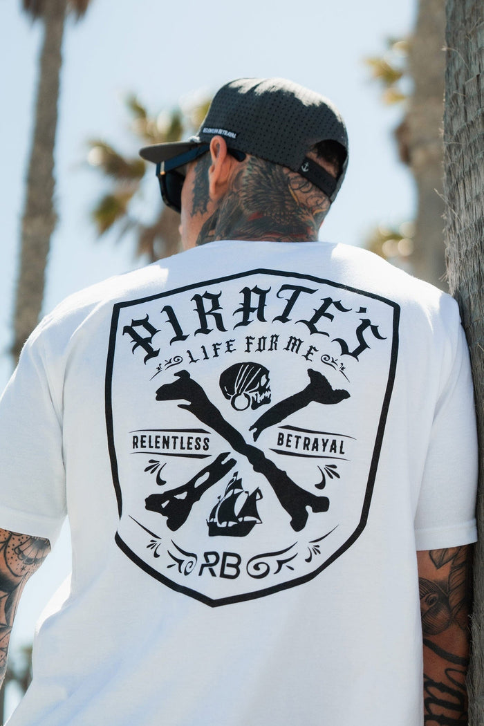 A tattooed person in sunglasses sports the Relentless Betrayal Pirate's Life White T-Shirt with bold black "Pirate's Life For Me," crossed bones, and nautical graphics on the back, standing by palm trees, embodying adventure.