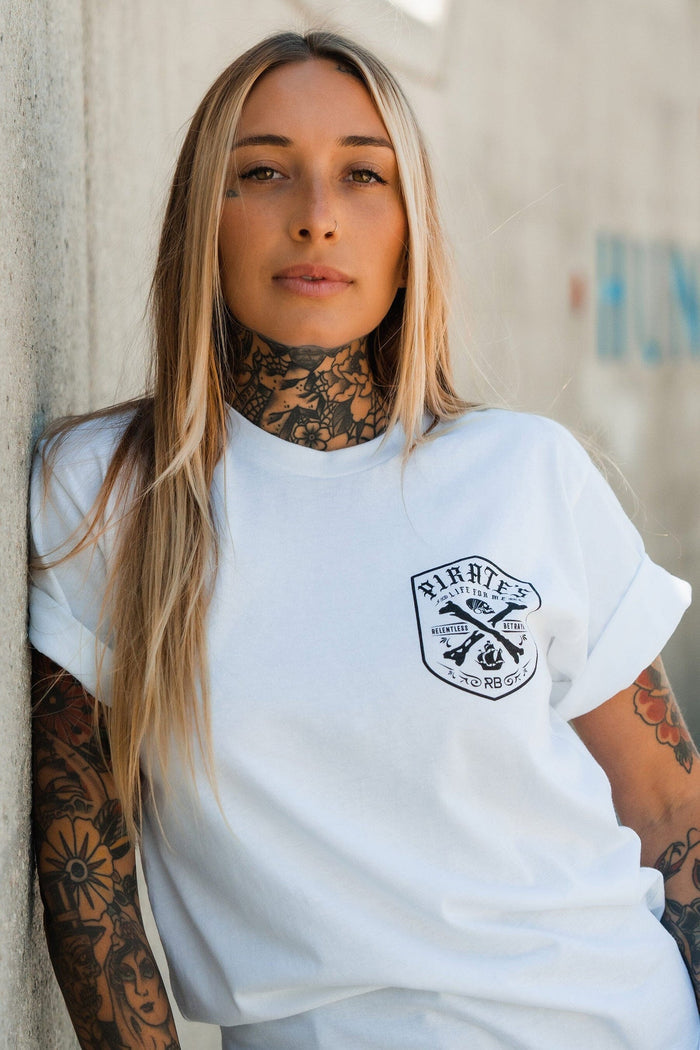 A tattooed woman with long blonde hair leans against a wall in the Relentless Betrayal Pirate's Life White T-Shirt, gazing confidently at the camera and channeling an adventurous spirit.