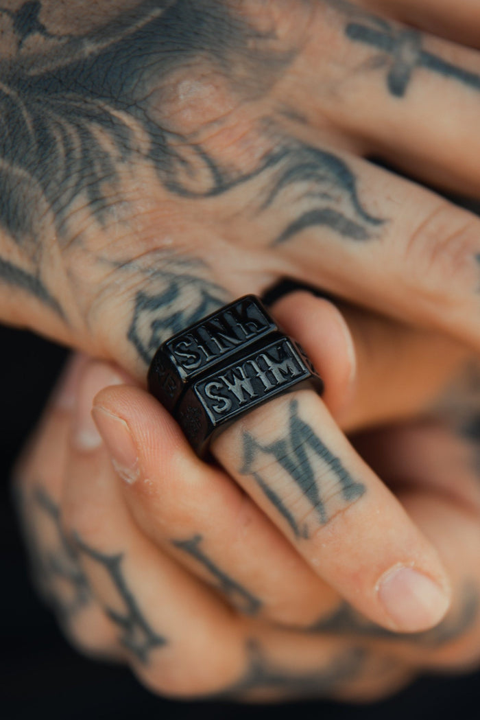 A tattooed hand adorned with Relentless Betrayals Polished Black Sink or Swim Rings, each engraved with SMILE, beautifully complements the intricate designs and numbers, echoing an ocean theme.