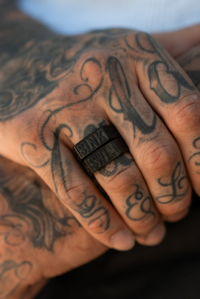 A hand adorned with intricate tattoos is showcased in close-up, highlighting a handcrafted ring from Relentless Betrayals Polished Black Sink or Swim collection. The blurred background emphasizes the tattoo artistry and striking stainless steel design of the ring.