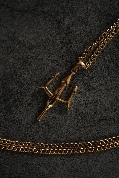A gold pendant from Relentless Betrayals Poseidons Trident Necklace shines on a dark, textured surface, with another chain partially visible beneath, embodying the maritime strength of Greek mythology jewelry.