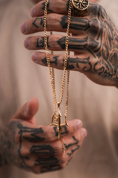 Tattooed hands clasp a Poseidons Trident Necklace by Relentless Betrayal, highlighting the maritime strength of Greek mythology jewelry. A ring with an intricate pattern adorns a finger against a softly blurred background, adding mystery.