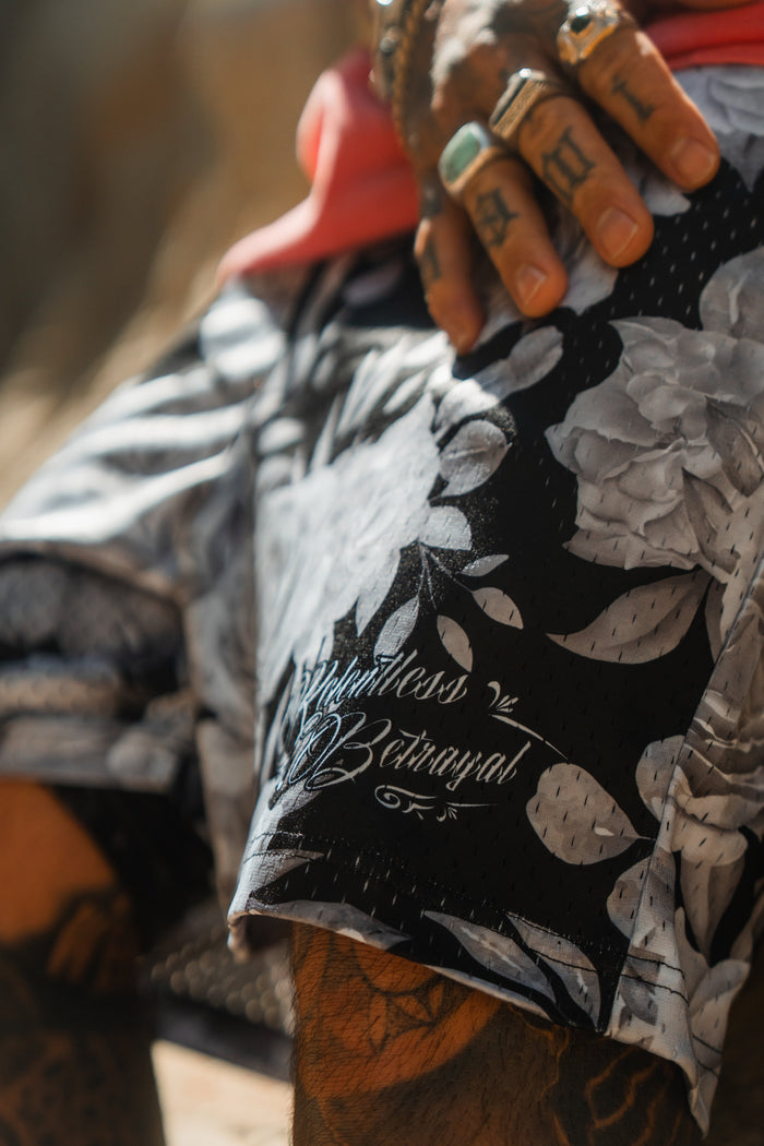 A tattooed hand with rings rests on Relentless Betrayal’s Premium Mesh Shorts | 5–Beauty in Darkness, featuring black and white floral patterns and cursive text. The unisex shorts are styled with a red shirt for a comfortable, bold look.