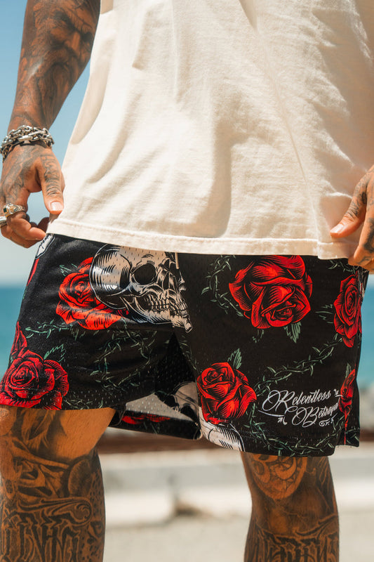 A person wearing Relentless Betrayal’s Premium Mesh Shorts | 5- Thorn Amongst Roses, featuring red rose and skull designs, stands by a body of water. Only their tattooed, jewelry-adorned lower half is visible.