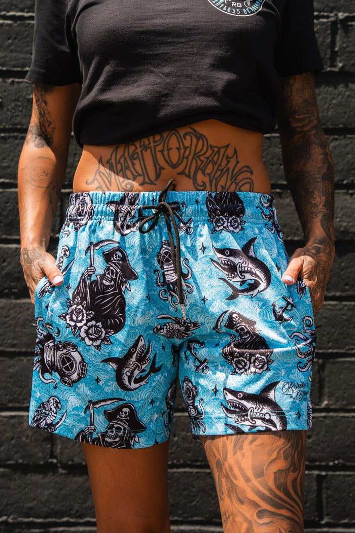 A person stands against a black brick wall, wearing Relentless Betrayal’s Premium Mesh Shorts | 5.5" - Davy Jones with black tattoo-style sharks, skulls, and roses. They have tattoos and a black cropped T-shirt.