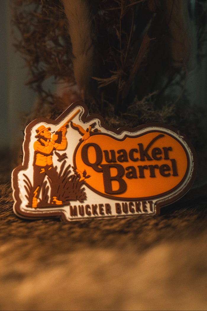 A decorative 3D PVC patch badge from Mucker Bucket, featuring an illustration of a hunter and the text "Quacker Barrel" at the bottom, displayed on a rustic surface. This Quacker Barrel PVC Patch measures 4x4 inches and comes with a Velcro backing for easy attachment.