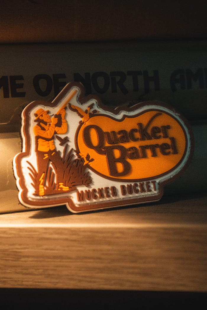 A 4x4 Quacker Barrel PVC Patch from Mucker Bucket features a hunter aiming a rifle, with text that reads "Quacker Barrel" and "Mucker Bucket," propped against books.