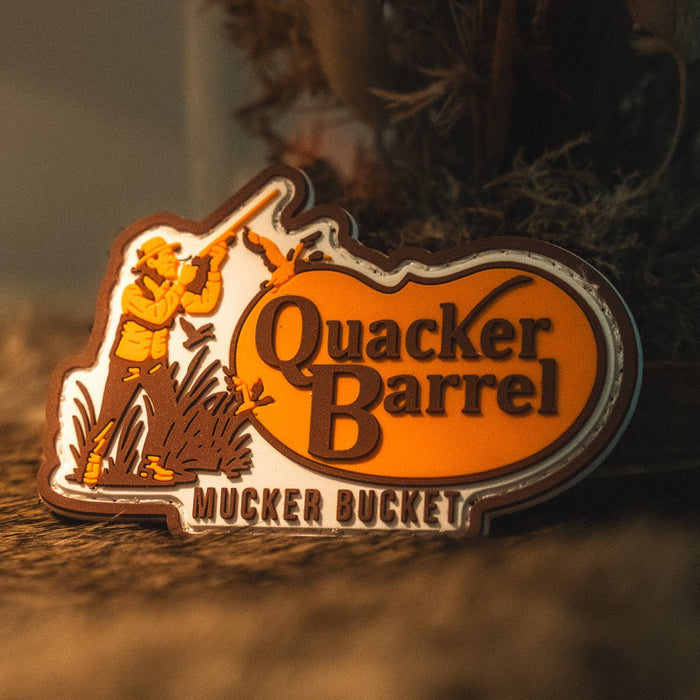 The Quacker Barrel PVC Patch by Mucker Bucket features a 3D design with the text "Quacker Barrel Mucker Bucket" alongside a cartoon man in a hat holding a shovel, set against a brown and orange background.