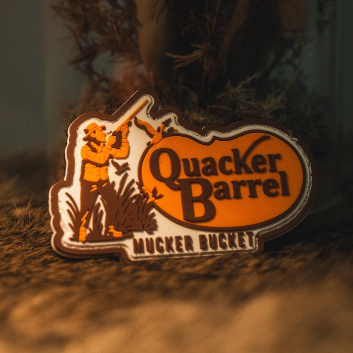 Introducing the Quacker Barrel PVC Patch by Mucker Bucket—a badge shaped like Kentucky, featuring the text "Quacker Barrel Mucker Bucket" and an illustration of a man aiming a gun in an orange and brown color scheme. This unique 3D PVC patch measures 4x4 inches and comes with Velcro backing for easy attachment.