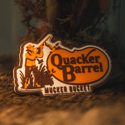 A Quacker Barrel PVC Patch by Mucker Bucket, featuring a person with birds and shaped like a state outline, rests on a textured brown surface—showcasing the bold style characteristic of 3D PVC patches.