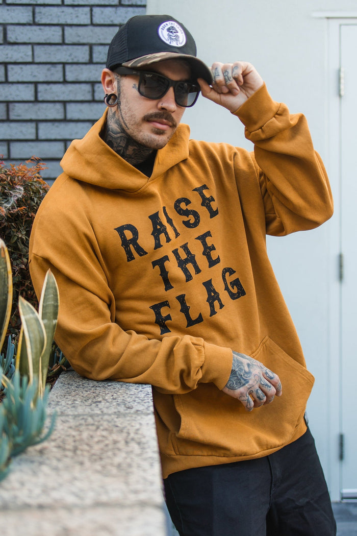 Wearing a Relentless Betrayal Raise the Flag Premium Toast Hoodie in mustard yellow, sunglasses, and a black cap, an individual stands outside by a plant-covered wall.