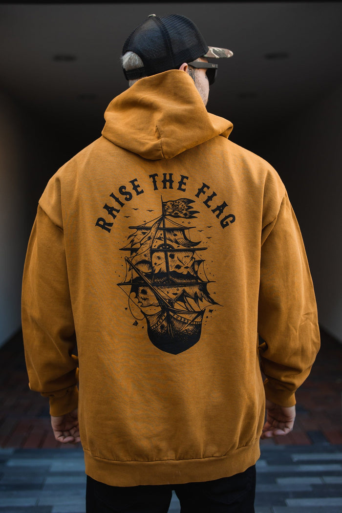 A person in a dim hallway wears the Raise the Flag Premium Toast Hoodie by Relentless Betrayal, featuring a black pirate ship design and RAISE THE FLAG text on the back. Theyre also wearing a baseball cap.