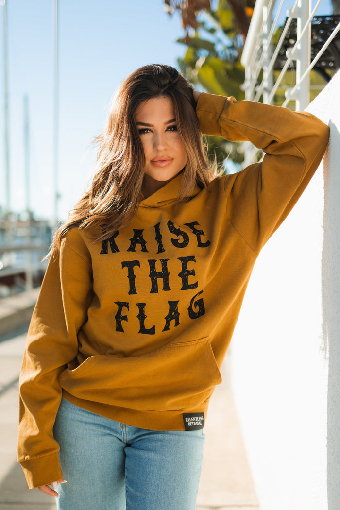 A person with long hair poses outdoors, resting one arm on a wall, wearing the Relentless Betrayals Raise the Flag Premium Toast Hoodie in mustard and light blue jeans. The blurred marina in the background evokes a pirate ships harbor.