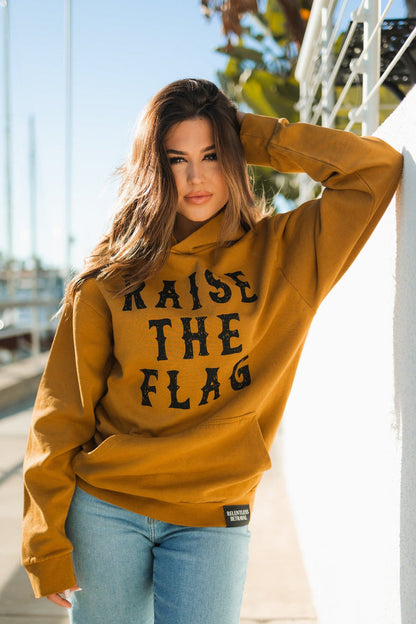 A person with long hair poses outdoors, resting one arm on a wall, wearing the Relentless Betrayals Raise the Flag Premium Toast Hoodie in mustard and light blue jeans. The blurred marina in the background evokes a pirate ships harbor.