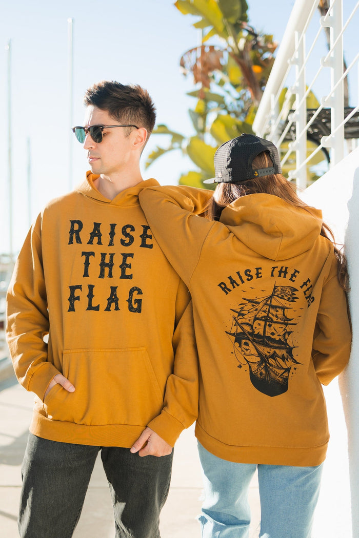 Two people wear matching Raise the Flag Premium Toast Hoodies from Relentless Betrayal. One features RAISE THE FLAG on the front, while the others back showcases a pirate ship design. They stand outdoors near a railing and palm trees.