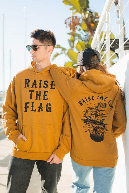 Two people wear matching Raise the Flag Premium Toast Hoodies from Relentless Betrayal. One features RAISE THE FLAG on the front, while the others back showcases a pirate ship design. They stand outdoors near a railing and palm trees.