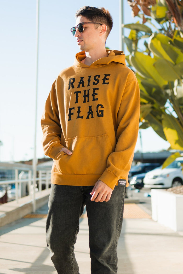 A person in a Raise the Flag Premium Toast Hoodie by Relentless Betrayal stands on a sunny sidewalk wearing sunglasses, with large green leaves and parked cars in the background, enhancing the relaxed vibe.