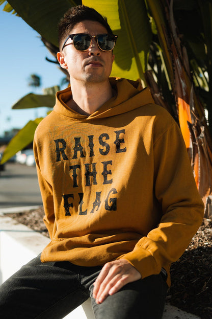 A person sits outdoors in a Raise the Flag Premium Toast Hoodie by Relentless Betrayal, sunglasses on, RAISE THE FLAG boldly displayed. Palm leaves sway under the sunny sky as visions of a pirate ship sailing on adventure drift through the breeze.