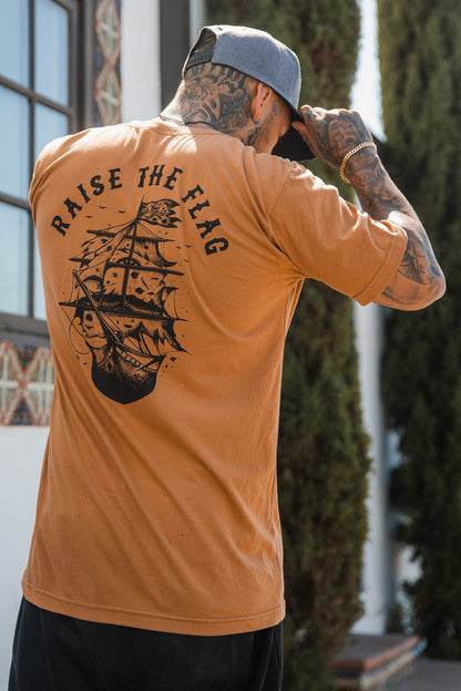 Near a tree and building, a tattooed individual adjusts their cap while wearing the Relentless Betrayals Raise the Flag Tee in brown jersey cotton. The T-shirt, adorned with a pirate ship and RAISE THE FLAG text on the back, captures a daring spirit of surrender or face an attack.
