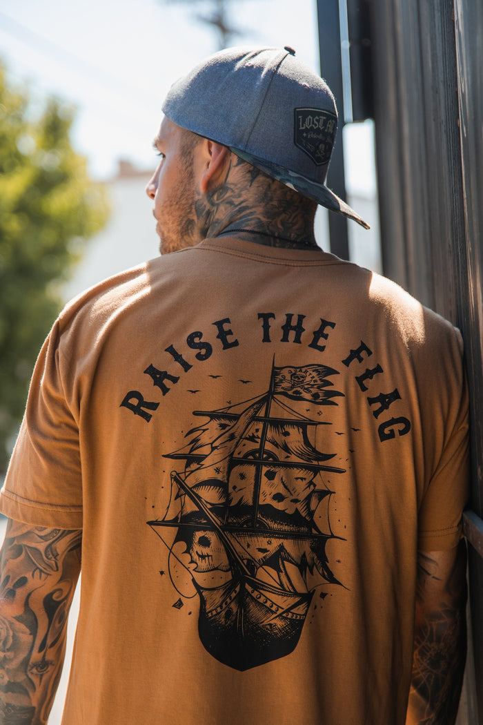 Outdoors by a building, a man in the Relentless Betrayals Raise the Flag Tee with ship and RAISE THE FLAG design, flaunts tattoos on his arms and neck. A backward baseball cap completes his look, evoking the bold spirit of an unfurled pirate flag.