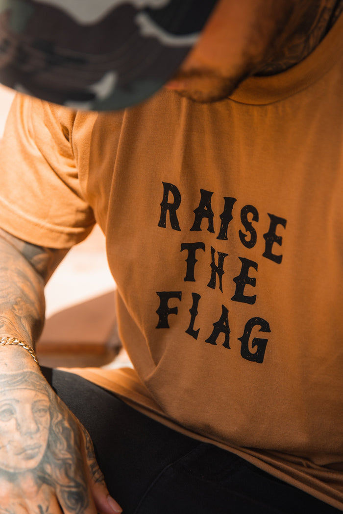 A person dons a Relentless Betrayal Raise the Flag Tee, reminiscent of a pirate flag, paired with a camouflage cap. Tattoos accentuate their arm, enhancing the rebellious look, with the focus on their upper body and striking shirt.