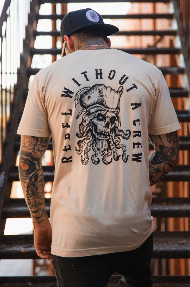 Wearing a cap and the unisex beige Rebel Without a Crew t-shirt by Relentless Betrayal, featuring a skull design and text on the back, a tattooed rebel descends metal stairs.