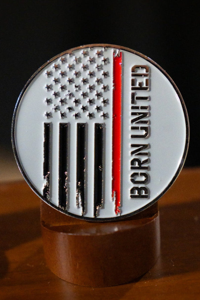 The Born United Private Label Red Line Flag Challenge Coin features a round design with black and white stripes, one bold red stripe for support, and "BORN UNITED" on the right side—a perfect fan collectible displayed on wood.
