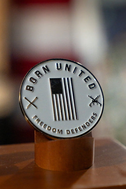 The Red Line Flag Challenge Coin by Born United Private Label features a stylized American flag, crossed rifles, and the words BORN UNITED and FREEDOM DEFENDERS—ideal fan gear displayed on a wooden stand.