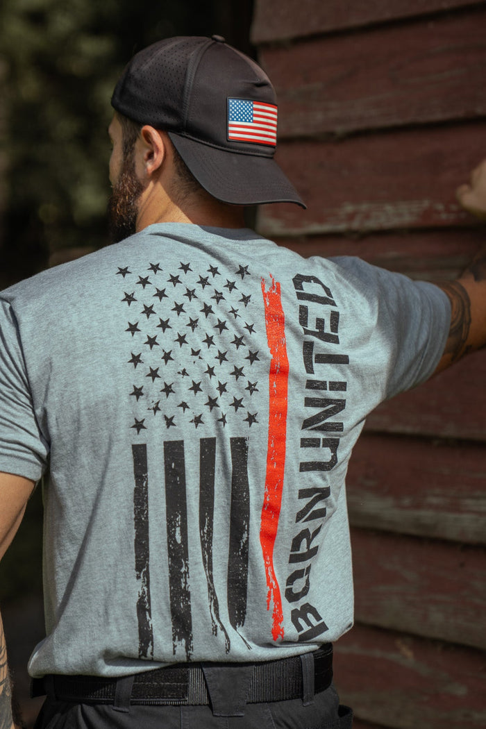 A man in a backward cap with a U.S. flag patch stands by a wooden wall, wearing the Born United Private Label Red Line Flag Tee featuring a distressed U.S. flag with a red stripe and BRAVERY printed beside it.
