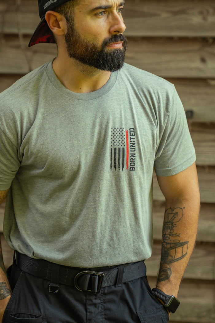 A bearded, tattooed man in a gray Red Line Flag Tee by Born United Private Label and a backwards black cap stands outdoors by a wooden wall, looking to the side.