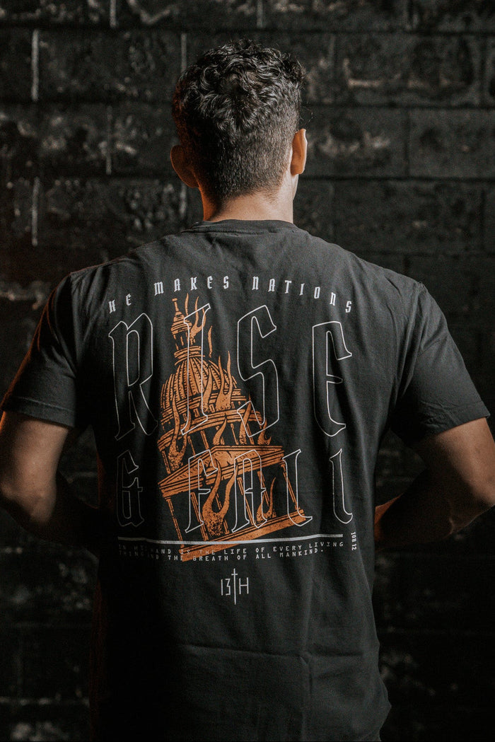 A person stands facing a dark, textured wall, wearing the Rise & Fall Premium Tee by 13th. The black T-shirt features an orange ship graphic and the words He Makes Nations Rise & Fall on the back. The dim lighting creates a dramatic mood, capturing the essence of lifes fleeting moments.