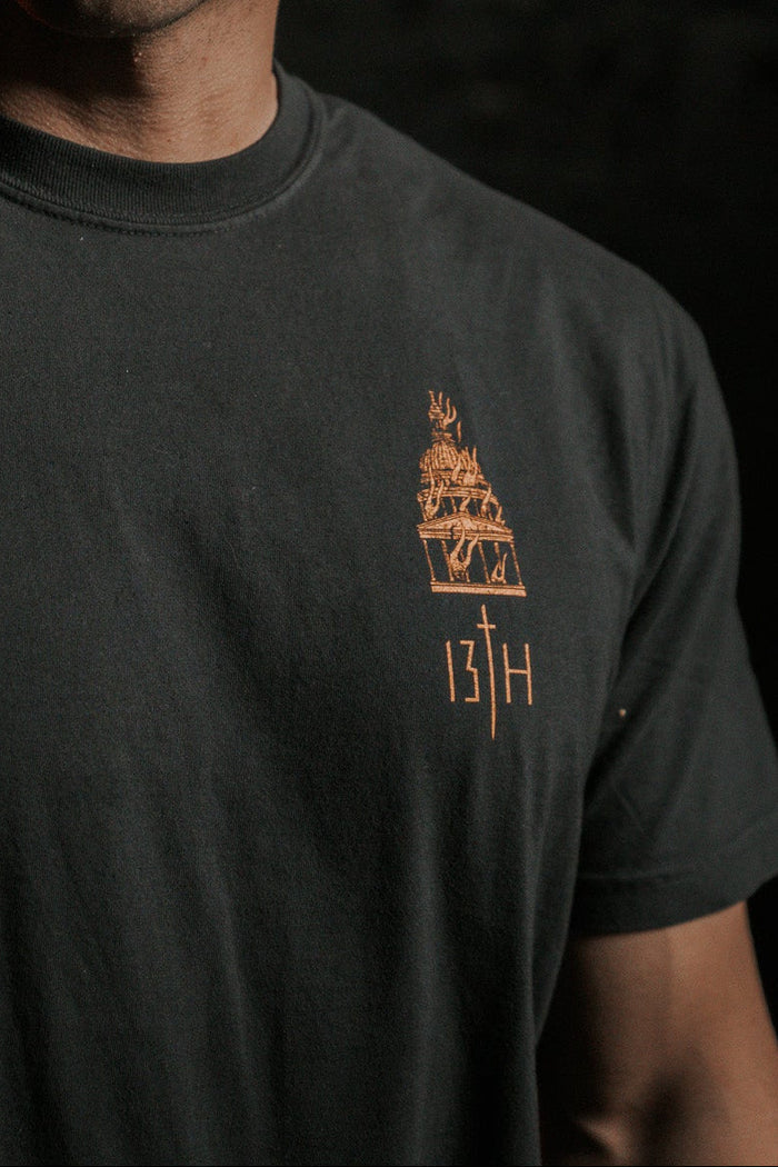 A man with short hair stands in a dimly lit room, embodying lifes essence. He wears the Rise & Fall Premium Tee by 13th, featuring a subtle design and the number 13H, symbolizing humanitys mysterious journey amidst a dark, shadowed background.