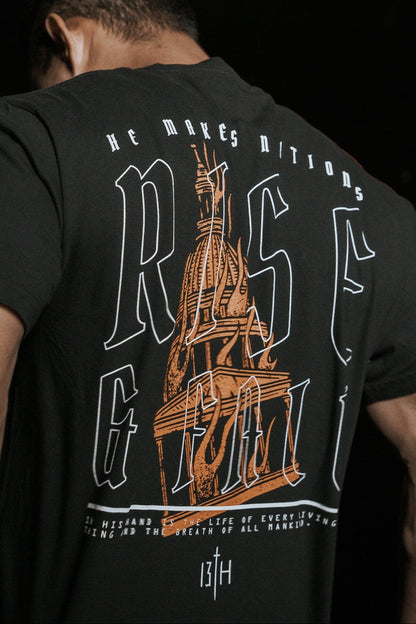 A person with short dark hair is seen from behind, wearing the black Rise & Fall Premium Tee by 13th. The T-shirt features large text RISE, a stylized building illustration, and small decorative text symbolizing mankinds journey.
