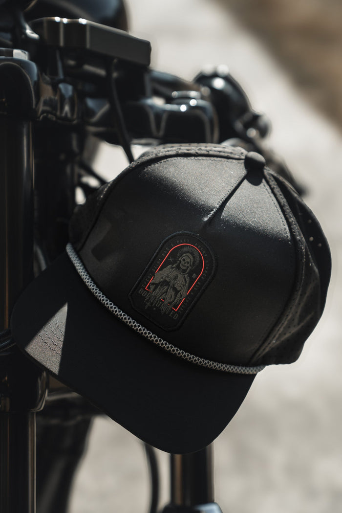 A Saints and Sinners Hat from Born United Private Label, featuring a Saint or Sinner? patch with a red outline of a woman, hangs on a motorcycle handlebar. Its textured band adds flair against the blurred outdoor setting, hinting at an adventure awaiting its owner.